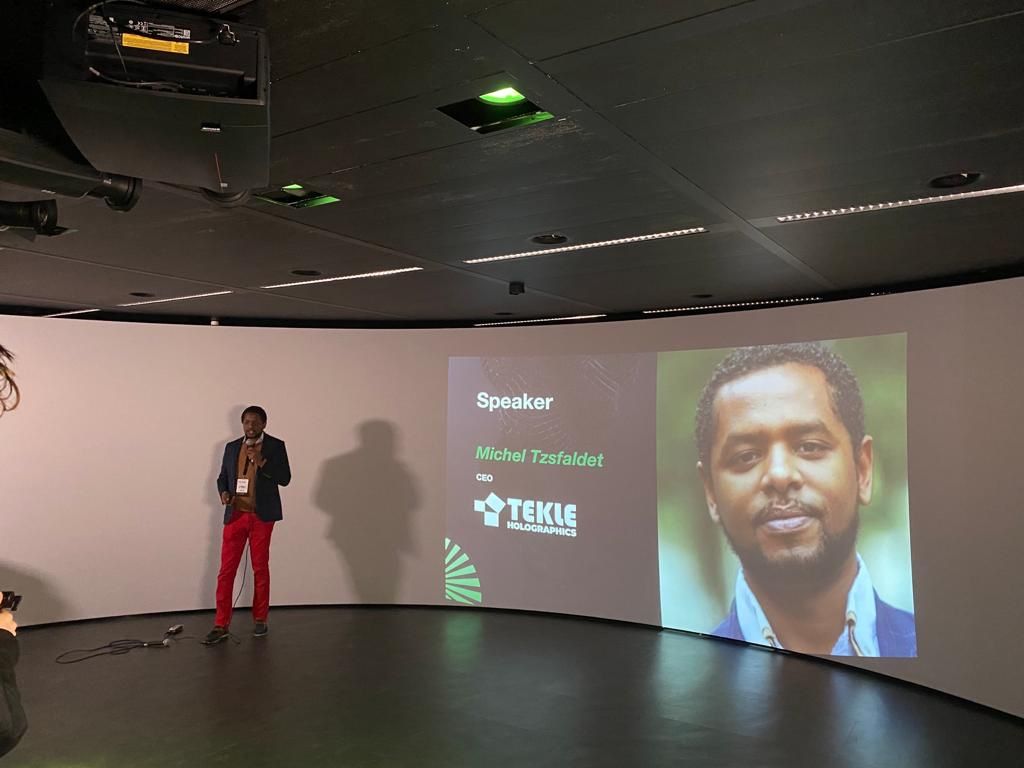 Michel Tzsfaldet on stage delivering a keynote in Amsterdam, his name and company shown on the screen behind him.