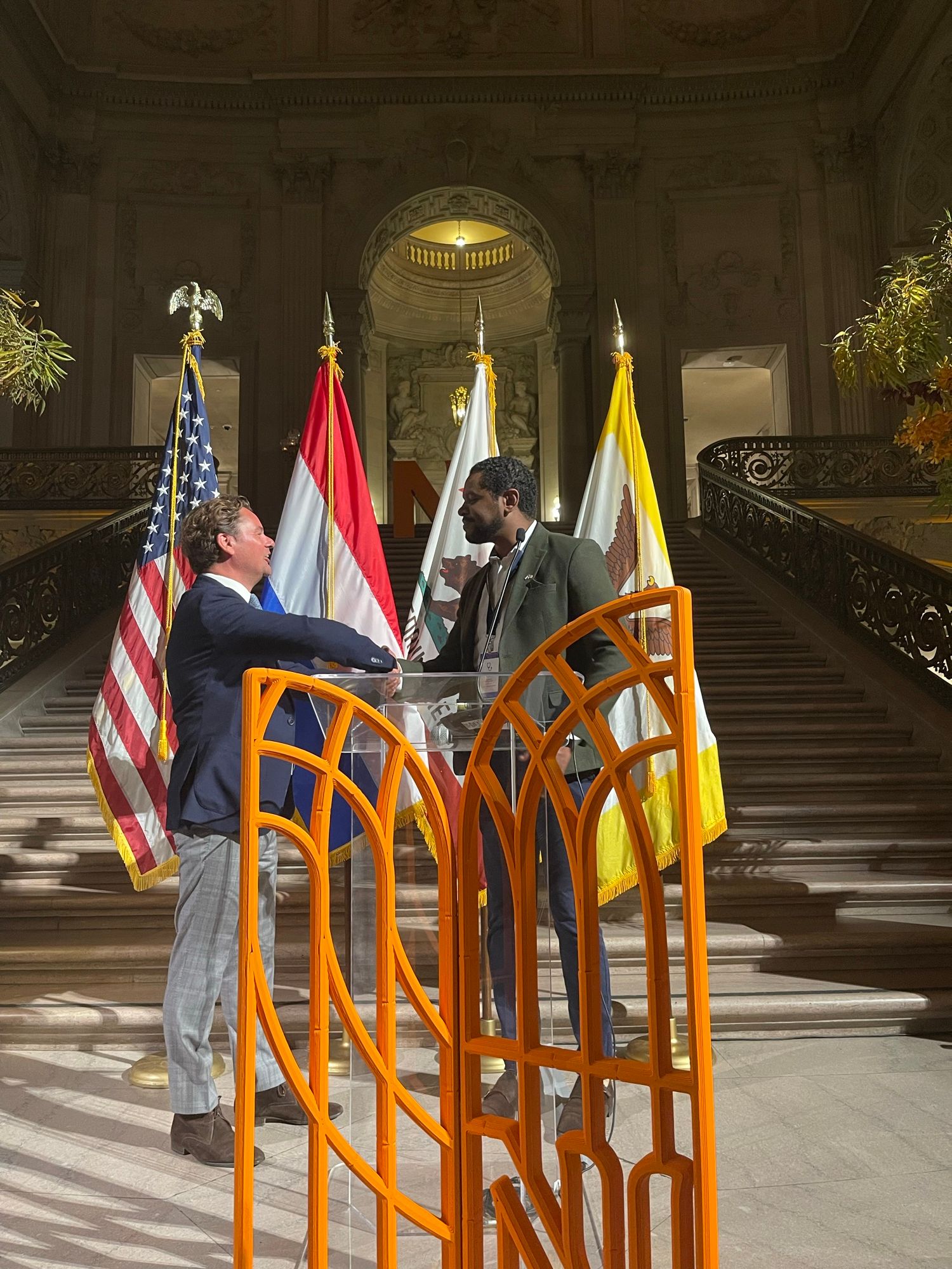 Michel Tzsfaldet shaking hands during the Netherlands economic mission to California, at San Francisco City Hall beside US and Dutch flags.